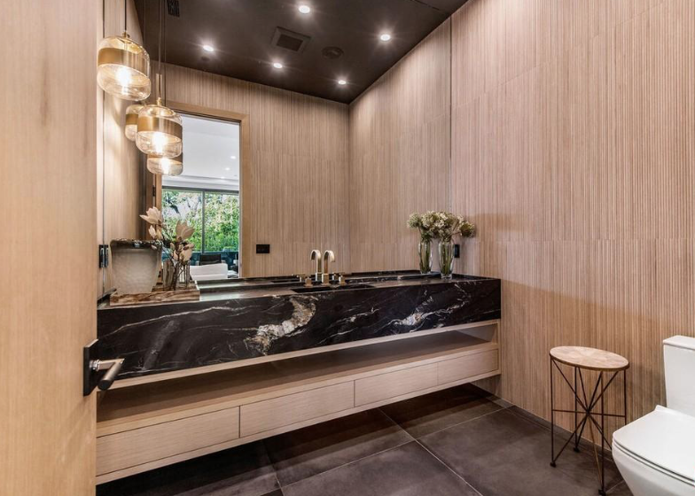 Powder room with black stone trough sink, light wood walls, and brass pendant lights.